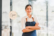 © kenchiro168 - Young Asian woman barista wearing apron standing with arms crossed in front of coffee shop entrance, smiling confidently, open sign hanging on glass door, modern cafe interior visible inside