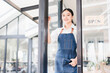 © kenchiro168 - Young Asian woman wearing denim apron standing at glass door of modern cafe, smiling and welcoming customers, bright natural light, urban background, friendly atmosphere