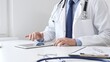 © rogerphoto - Male doctor wearing lab coat and stethoscope using digital tablet while sitting at desk in medical office, with medical chart and pen in the foreground