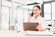 © kenchiro168 - Young woman business attire sitting at desk holding tablet talking on smartphone smiling modern office natural light professional confident productive