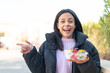 © luismolinero - Young woman at outdoors holding a bowl of fruit at outdoors surprised and pointing finger to the side