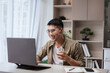 © ME Image - Asian man smiling while working from home, wearing glasses and wireless earbuds, holding coffee cup during video call on laptop. Perfect for remote work, online meetings, and freelance lifestyle