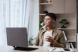 © ME Image - Asian man wearing glasses and earbuds holding coffee cup, looking thoughtful while working from home at laptop. Modern home office concept for telework, focus, and digital productivity themes