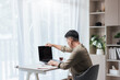 © ME Image - Asian man at a home desk opens his laptop to start work. A notebook, pen, and small cup sit beside him. Daylight through sheer curtains, with shelves and indoor plants in a clean, minimalist room.
