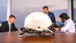 © Muhamad - A white hard hat rests on a table in the foreground, with blurred businessmen in the background