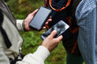 © DragonImages - Young adult Caucasian man holding smartphone and portable solar power bank outdoors next to another , using devices while hiking or exploring forest environment