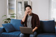 © Crystal - Young asian man suffering from eye strain while working remotely from home, holding his laptop on the sofa and rubbing his tired eyes