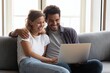 © saha - Happy young couple sitting on gray sofa laughing while looking at laptop together in bright living room, enjoying relaxed and joyful moment at home