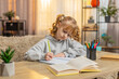 © Andrii Iemelianenko - Young school girl sitting at table on sofa, focused on solving a task in notebook. Caucasian child kid happily celebrating success, showing enthusiasm and joy during homework at home. Education study