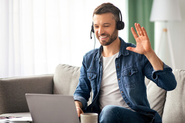 Smiling bearded businessman having video conference from home, using headset and laptop, talking and gesturing. Cheerful middle-aged man sitting on couch in living room, having video chat, copy space