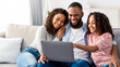 © Prostock-studio - Precious Time With Family. Portrait of happy afro parents and cute little girl using laptop together, watching photos or movie, browsing internet, choosing gifts online sitting on sofa in living room