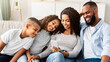 © Prostock-studio - Portrait of happy black family spending time together, woman holding and using smartphone, sharing media sitting on the sofa in living room. Mom and dad watching video or photos with children