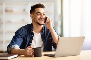 Happy arab guy freelancer working from home, having phone conversation with client or boss, sitting at workdesk in front of laptop, drinking coffee, looking at copy space and smiling