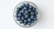 © Lacobus - Overhead shot of fresh blueberries in a clear glass bowl on a white surface in bright light