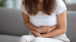 © oksa_studio - Close up of woman sitting on a sofa at home, holding her belly and experiencing discomfort from abdominal pain or stomach ache