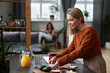 © TrueFrame Collective - Horizontal medium portrait shot of joyful young woman with blond hair cooking breakfast while her girlfriend relaxing on sofa surfing internet