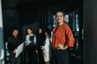 © qunica.com - A confident woman in a red blouse stands in a modern office, leading a team meeting. The diverse group of employees collaborates in a professional setting, showcasing teamwork and leadership.