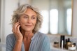 © SerPak - A mature woman with curly gray hair touching her ear, looking thoughtfully in a bright bathroom with cosmetics in the background.