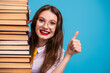 © deagreez - Smart woman with stack of book wearing glasses and showing thumbs up against bright blue background in studio