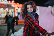 © qunica.com - A cheerful young woman in a red coat poses at a nighttime outdoor winter event, surrounded by festive lights and activity.