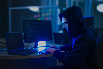 A mysterious hooded hacker working on a computer in a dark room, illuminated by blue screen light. A concept for cybersecurity, hacking, cybercrime, and data privacy threats.