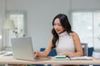 © amnaj - Asian businesswoman working with laptop computer, holding pen and looking at screen, sitting at desk in modern office with documents and notebooks