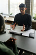 © pressmaster - Vertical shot of serious young Hispanic man sitting at desk in front of his Black co-worker discussing new business plan