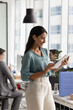 © fizkes - Brazilian professional woman working online using digital tablet standing in contemporary coworking office with colleagues in background. New AI application or business tools usage, workflow remotely