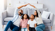 © Prostock-studio - Housing For Family Concept. Portrait of positive black man and woman making symbolic roof of hands above their cheerful little son and daughter, looking up, sitting on floor carpet in living room