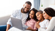 © Prostock-studio - Portrait of cheerful African American parents and their happy children using laptop on pillow, watching movie or having online video conference, sitting on the couch in living room at home together