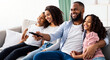 © Prostock-studio - Comfort And Relax. Portrait of happy loving black family of four people watching tv, smiling parents embracing their two children sitting on couch in the living room, dad holding remote control