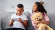 © Prostock-studio - Children Entertainment And Leisure. Portrait of excited black boy playing online mobile game on his smartphone, interested girl looking at the dagdet screen, hugging teddy bear, sitting on couch
