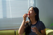 © amnaj - Asian woman taking pill, holding glass of water, sitting on sofa at home, sunlight through blinds