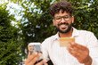 © Maria Vitkovska - Smiling young man making online payment using credit card and smartphone outdoors