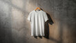 © karthik - Minimal white blank T‑shirt mockup on wooden hanger against textured concrete wall with dramatic studio lighting and long shadows, perfect for apparel branding, print design, and e‑commerce product pr