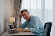 © Tj - Senior man using laptop computer working from home office smiling and looking at the screen, he's wearing a blue shirt sitting at the desk with a lamp and a coffee cup
