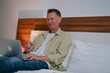 © Tj - Smiling mature man using laptop sitting comfortably in bed in hotel room, enjoying online communication or remote work during business trip or vacation