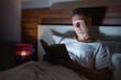 © Tj - Middle-aged man enjoying a book under the covers in his bed at night, illuminated by a bedside lamp, finding peace and relaxation before sleep