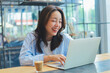 © Mallika - Cheerful Asian woman smiling and laughing while using laptop at modern cafe, representing freelance lifestyle, digital nomad, and online business success.