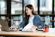 © Natthawadee - A young woman sits at a desk with a laptop, writing notes on documents. She works in a bright office environment, focused, productive, and engaged in her tasks.