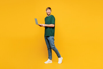 Full body side view young IT man he wear green t-shirt casual clothes hold use work on laptop pc computer walking going isolated on plain yellow orange background studio portrait. Lifestyle concept.
