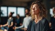 © Tina - A group of people meditating in a serene indoor environment. A young Caucasian woman with curly hair sits in front, eyes closed, while diverse individuals are visible in the background.