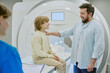 © AnnaStills - Caucasian child sitting on MRI scanner smiling at bearded Caucasian man standing beside, medical professional holding clipboard in foreground, hospital diagnostic setting