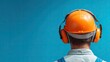 © denish - Rear view of a construction worker wearing a hard hat and earmuffs against a blue wall