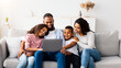 © Prostock-studio - Family Weekend. Portrair of cheerful African American parents and their little children using laptop at home together, watching movie or browsing internet, sitting on the couch in living room