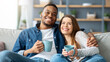 © Prostock-studio - Home Leisure. Cheerful interracial couple watching tv and drinking coffee on couch, romantic young black man and white woman relaxing in cozy living room, enjoying domestic pastime, free space