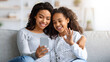 © Prostock-studio - Cheerful beautiful black mother and daughter having video call, sitting on couch and using modern smartphone, waving and smiling at mobile phone screen, living room interior, copy space