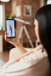 © DC Studio - Asian female freelancer seated on couch at home, holding smartphone during online consultation with male physician. Remote medical session with healthcare professional in progress.