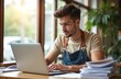 © Viktor - Young man in apron works on laptop near stacks of bills, documents. Small business owner manages finances, bookkeeping, payments, taxes. Busy cafe employee multitasking with computer for restaurant