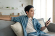 © Monster Ztudio - Smiling handsome young Asian man sitting on cozy couch, using funny mobile apps in living room. man at home, doing online shopping, messaging friends, posting on social media